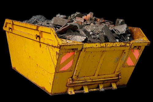 A yellow industrial skip filled with broken concrete, brick fragments, and assorted rubble, positioned outdoors on a paved surface. The skip's surface displays signs of wear with visible dirt and scratches, and it features two reflective orange and pink safety markers on the rear side. The rubble inside appears to be a mixture of rough-textured concrete pieces with jagged edges and reddish-brown brick debris, with some pieces protruding above the rim of the skip. The background is entirely black, emphasizing the skip and its contents, suggesting the image was taken in a controlled studio or isolated setting. This scene is relevant to waste management and rubbish removal services offered by Commercial Waste Removal Tottenham, highlighting the type of construction debris typically collected and disposed of in the Tottenham area.