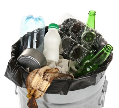 A close-up view of a filled black rubbish bin lined with a plastic bag, containing various discarded items such as a crumpled black plastic bag, a metallic tin can, a white plastic bottle with a green cap, and two green glass bottles, all related to household waste. The bin is situated outdoors on a paved surface, with some wooden debris and paper wrapping visible among the rubbish. The background is plain and white, focusing attention entirely on the heap of waste inside the bin, which illustrates typical refuse collected during commercial or residential rubbish removal services in Tottenham. The assorted materials display different textures, with smooth plastic, shiny metallic surfaces, and rough, weathered wood, reflecting the variety of waste that companies like Commercial Waste Removal Tottenham handle regularly. The overall scene emphasizes the importance of proper waste disposal and collection, consistent with local rubbish removal operations in the Tottenham area, helping to maintain cleanliness and environmental standards.