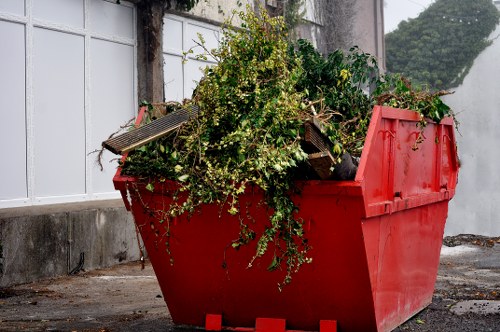 A large red plastic skip bin filled with green garden waste, including branches, leaves, and small twigs, positioned on a paved area near a building with white-framed windows. The waste in the bin appears to be organic and biodegradable, with some loose foliage cascading over the sides. The surrounding environment includes a concrete base and a grey wall, with a partial view of a hedge or shrubbery in the background. The scene suggests a commercial or domestic rubbish removal service associated with companies like Commercial Waste Removal Tottenham, serving areas such as Tottenham or surrounding postcode districts, involving garden clearance or general waste disposal. The lighting indicates an outdoor daytime setting with natural light illuminating the scene, emphasizing the vibrant red of the bin and the green hues of the waste materials, consistent with typical rubbish removal activities in London suburbs.